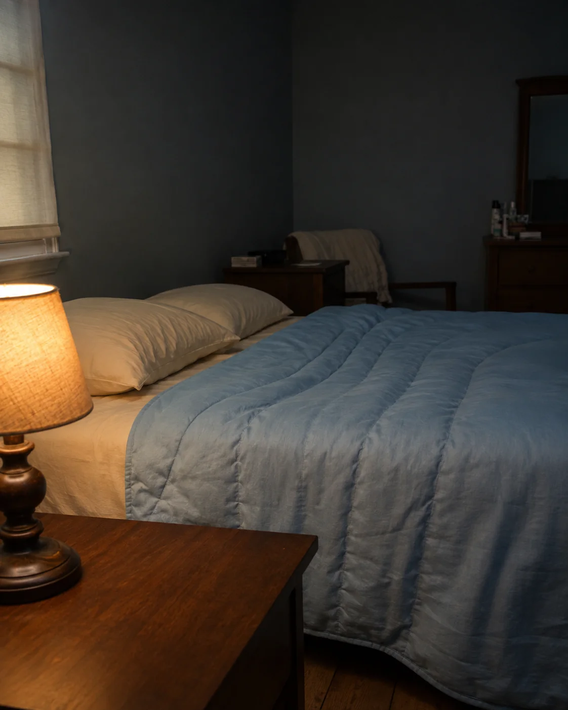 The blanket across the bed seen past a bedside lamp in the evening.